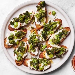 Assembled toasts smeared with ricotta and topped with broccoli on platter beside a small bowl of hot honey for drizzling...