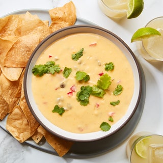 A bowl of hatch chile con queso being served with tortilla chips and margaritas.