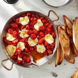 Overhead view of a baking dish with blistered tomatoes and goat cheese alongside toasted baguette.